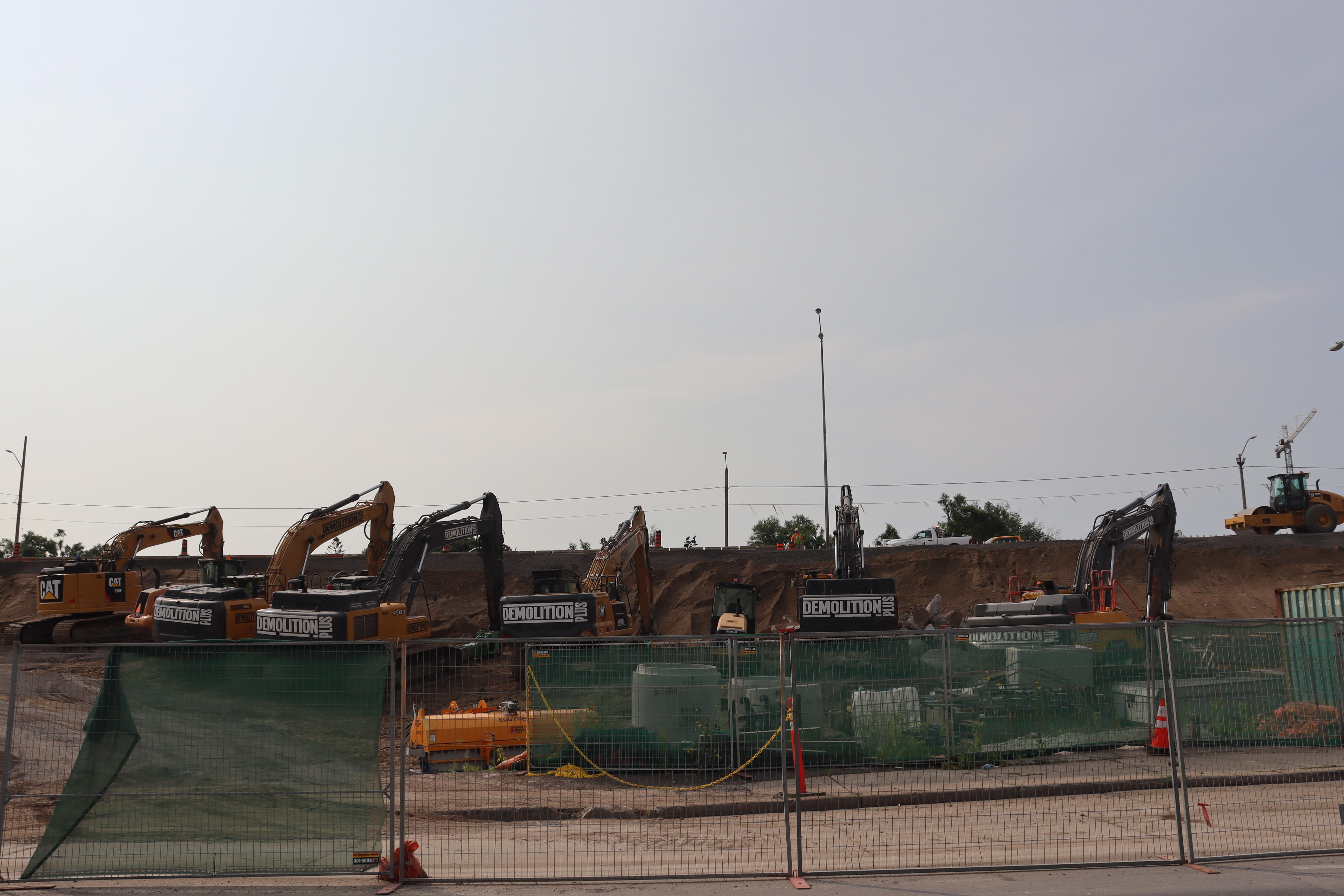 A row of excavators and other construction machinery, behind a temporary fence.